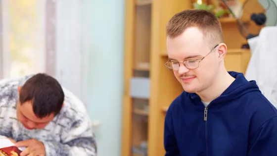 Young man looks at his work in a library setting alongside classmate.